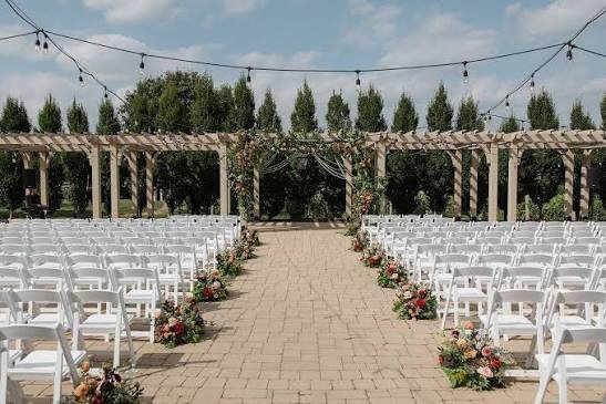 The pergola ceremony space at Bourgmont: a brick aisle, white chairs, a floral arch, and string lights overhead.